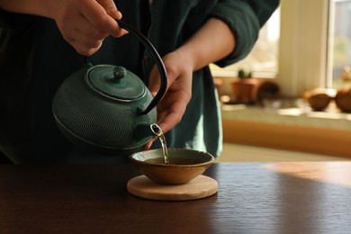 Photo of Woman pouring freshly brewed tea from teapot into cup at wooden table indoors, closeup and space for text. Traditional ceremony