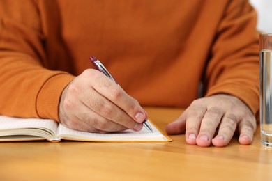 Photo of Young man writing in notebook at wooden table, closeup