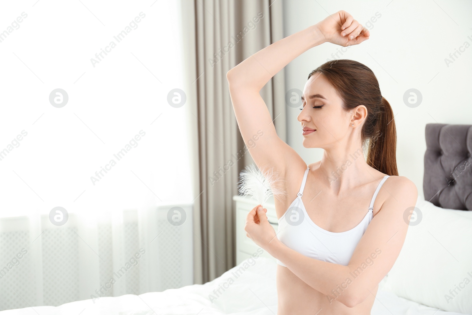 Photo of Young woman touching armpit with feather in bedroom, space for text. Epilation procedure
