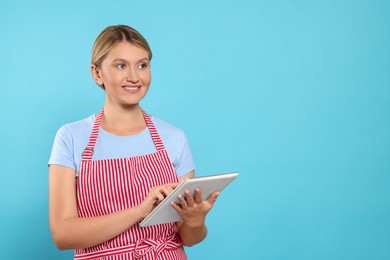 Photo of Beautiful young woman in clean striped apron with tablet on light blue background. Space for text