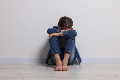 Photo of Child abuse. Upset girl sitting on floor near white wall