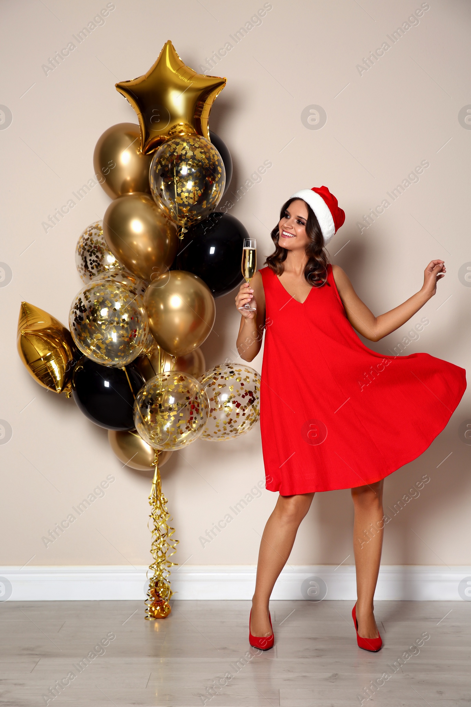 Photo of Happy woman in Santa hat with champagne and air balloons near beige wall indoors. Christmas party