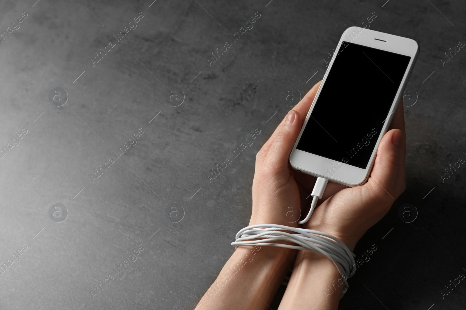 Photo of Woman holding mobile phone in hands tied with charging cable on grey background, closeup. Loneliness concept