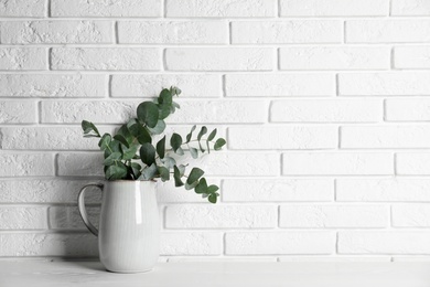 Photo of Beautiful eucalyptus branches in vase on white wooden table near brick wall. Space for text