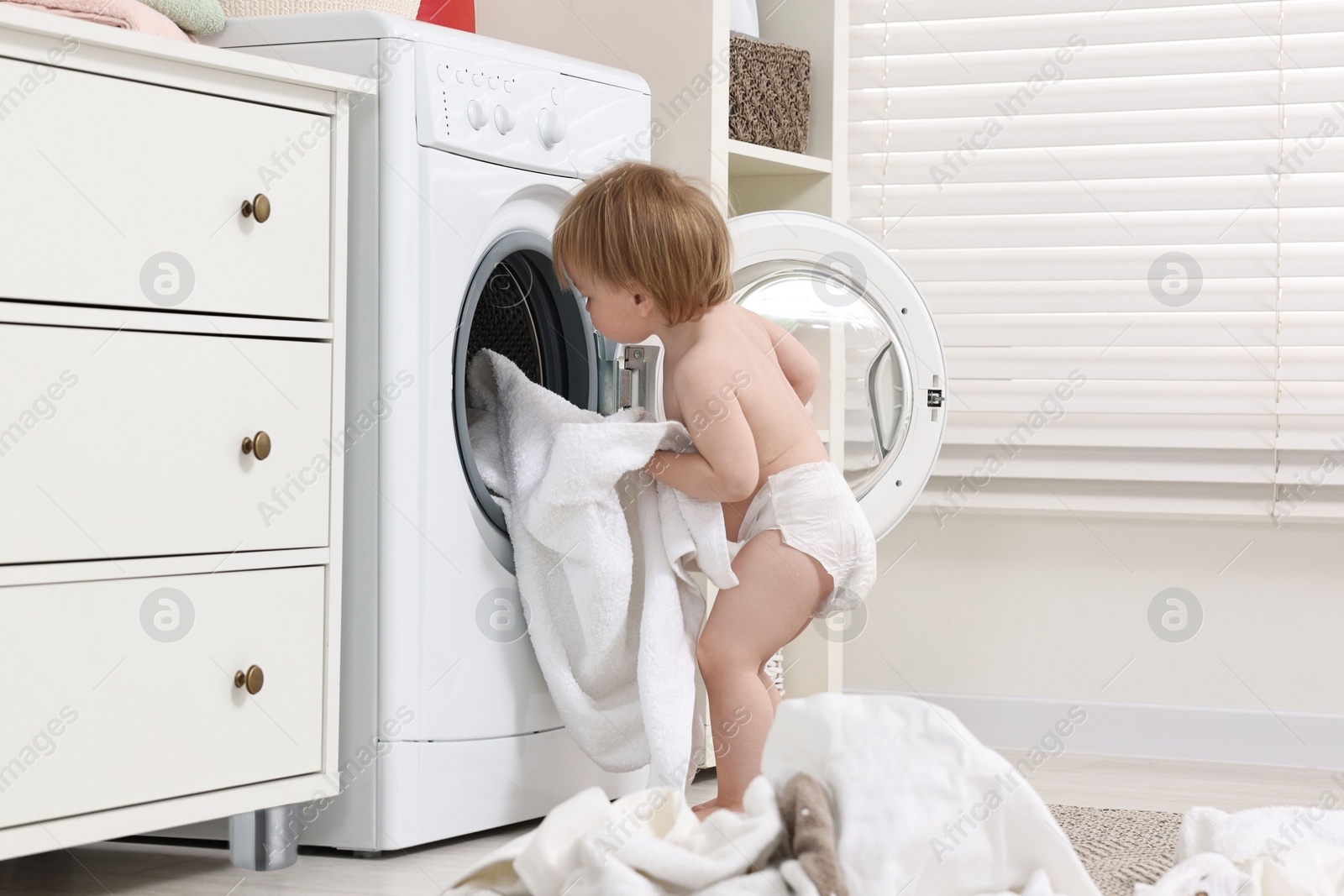 Photo of Little girl pulling baby clothes out of washing machine indoors