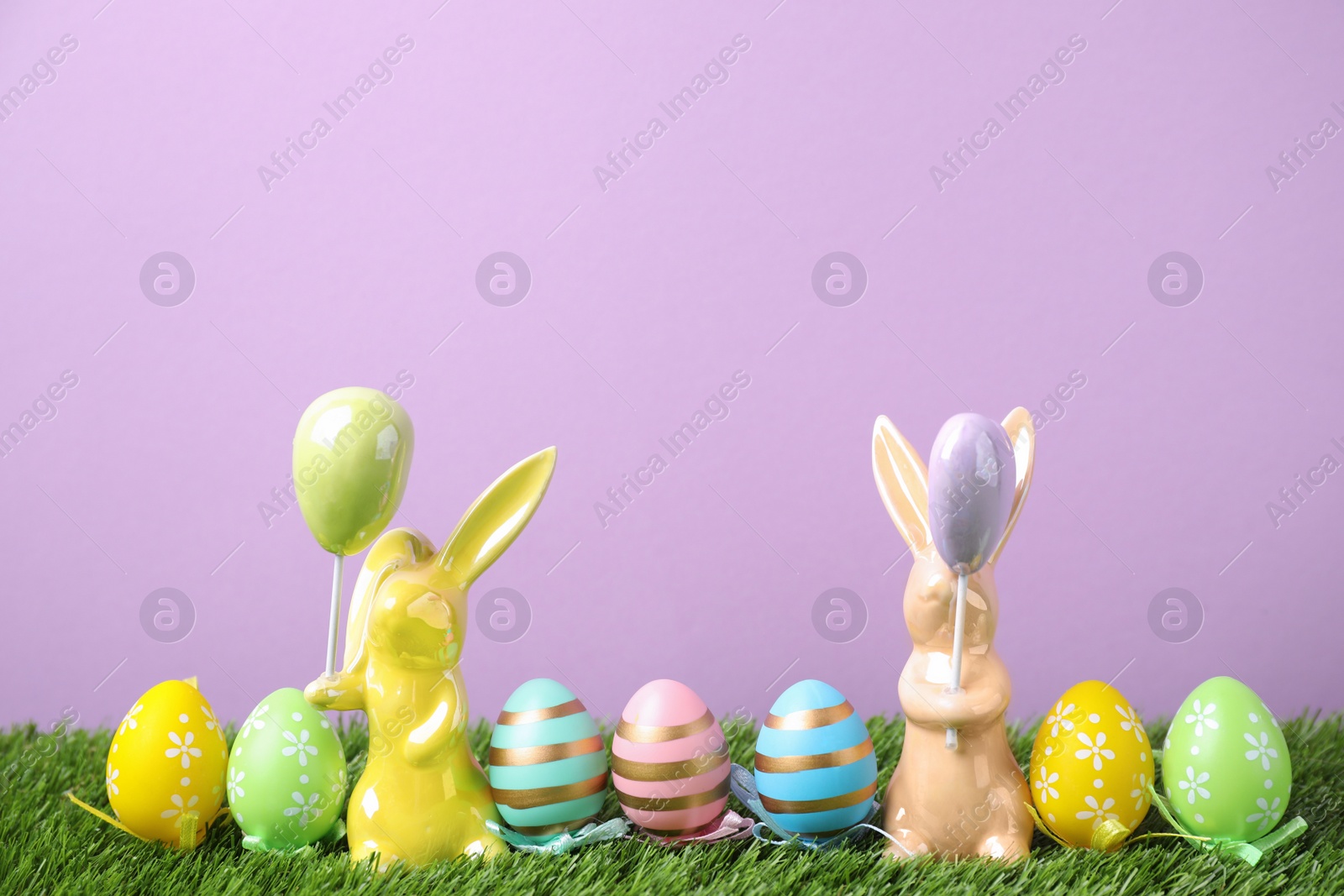 Photo of Easter bunny figures and dyed eggs on green grass against violet background