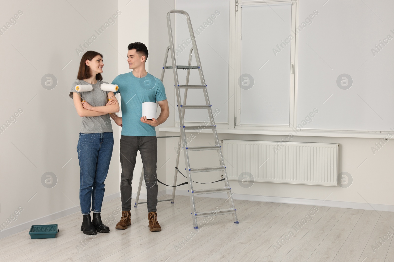 Photo of Young couple with painting tools near metal stepladder indoors. Room renovation