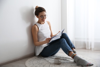 Photo of Young woman with cup of coffee reading book on floor at home