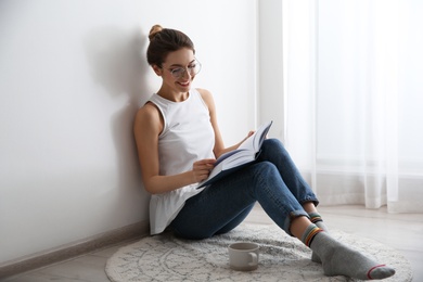 Young woman with cup of coffee reading book on floor at home