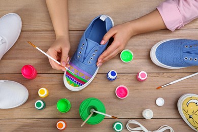 Photo of Woman painting on sneaker at wooden table, top view. Customized shoes