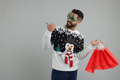 Photo of Happy young man in Christmas sweater and funny glasses with shopping bags on grey background. Space for text