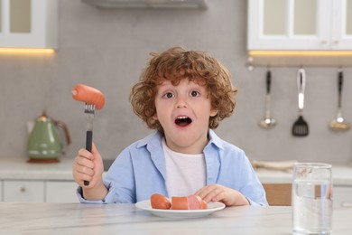 Photo of Cute little boy holding fork with sausages at table in kitchen