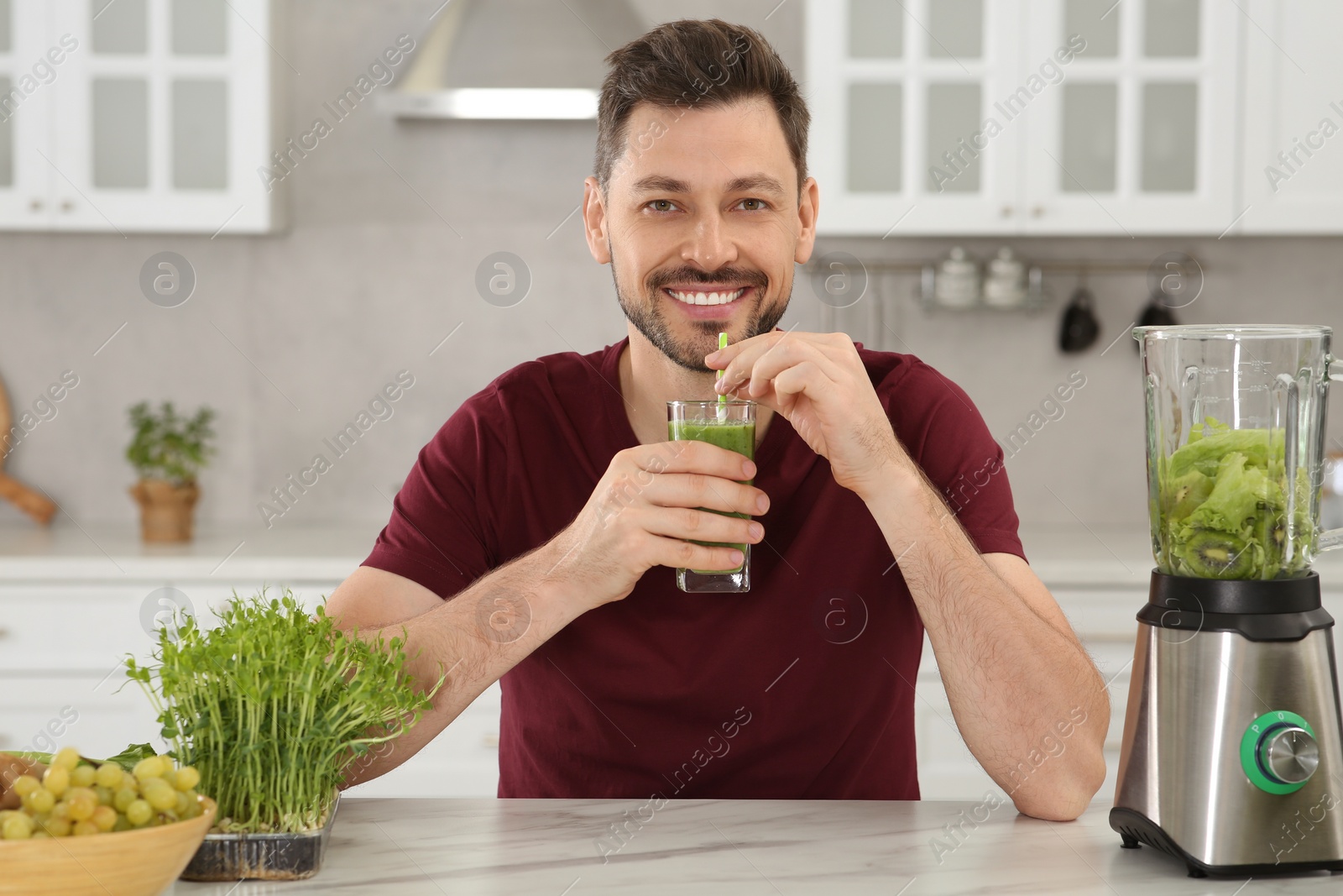 Photo of Happy man drinking delicious smoothie at white marble table in kitchen