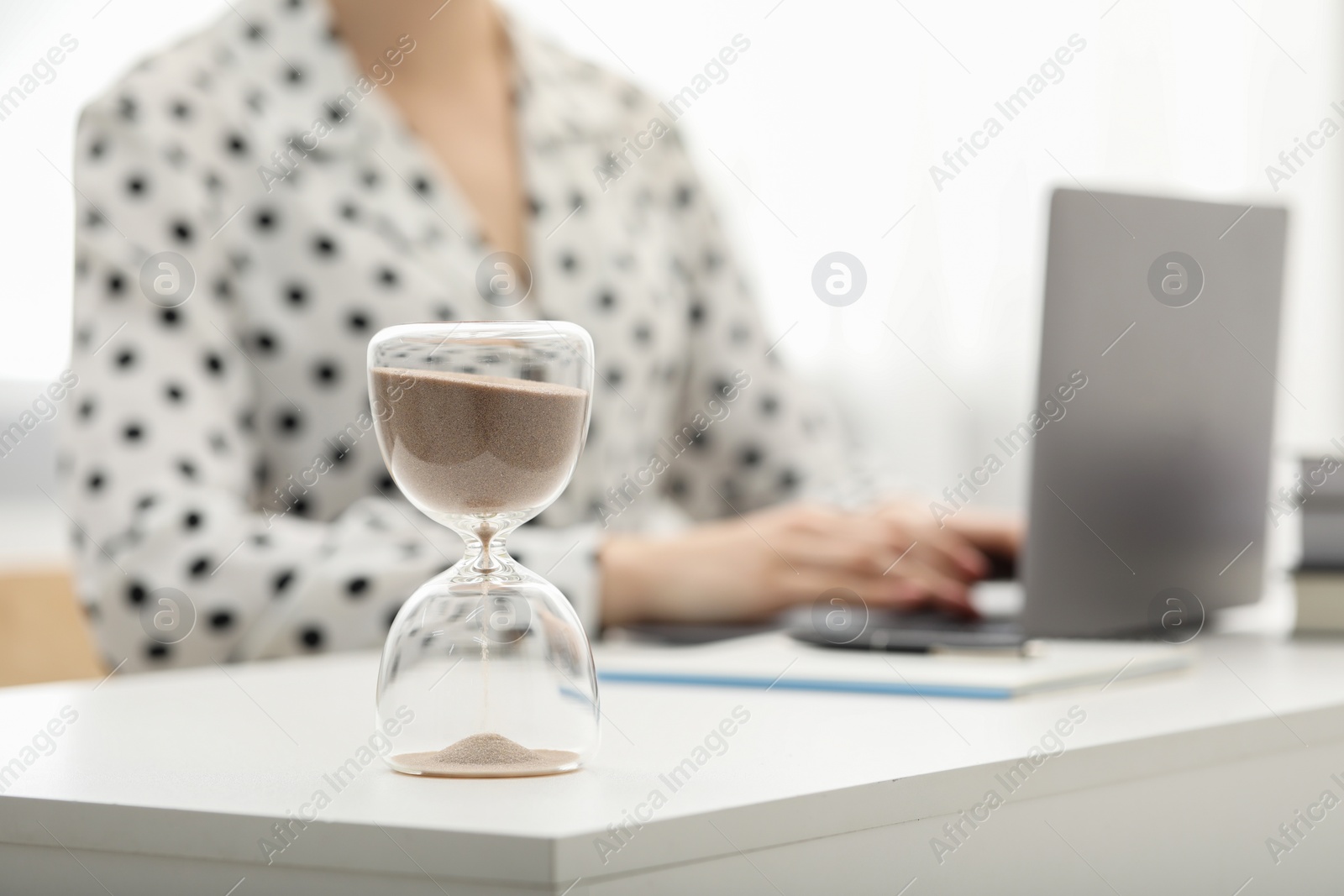 Photo of Hourglass with flowing sand on desk. Woman using laptop indoors, selective focus