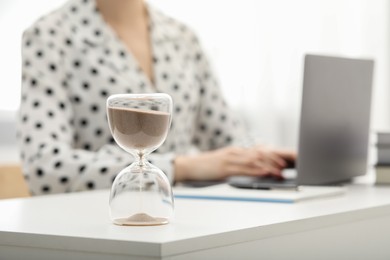 Photo of Hourglass with flowing sand on desk. Woman using laptop indoors, selective focus