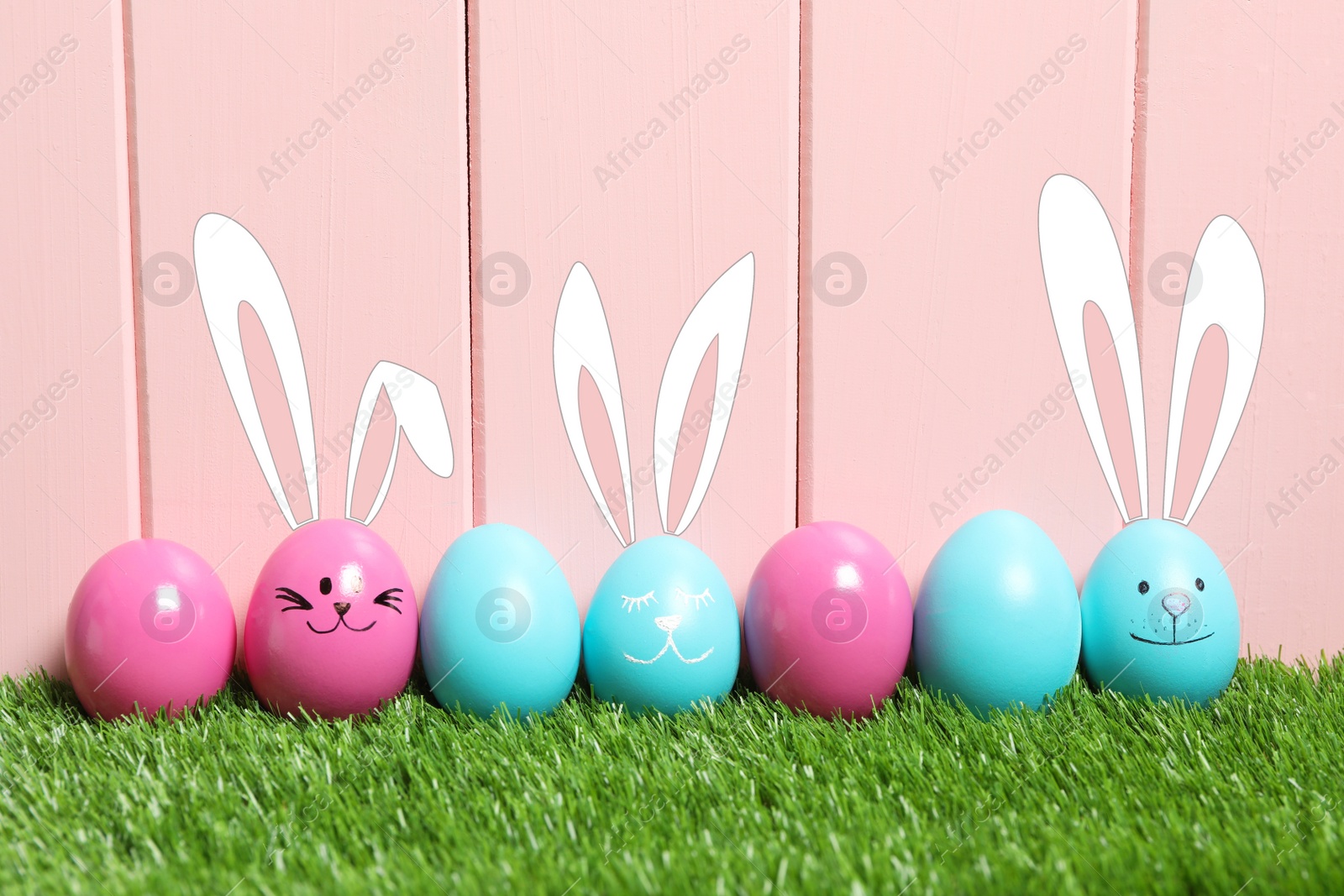 Image of Several eggs with drawn faces and ears as Easter bunnies among others on green grass against pink wooden background