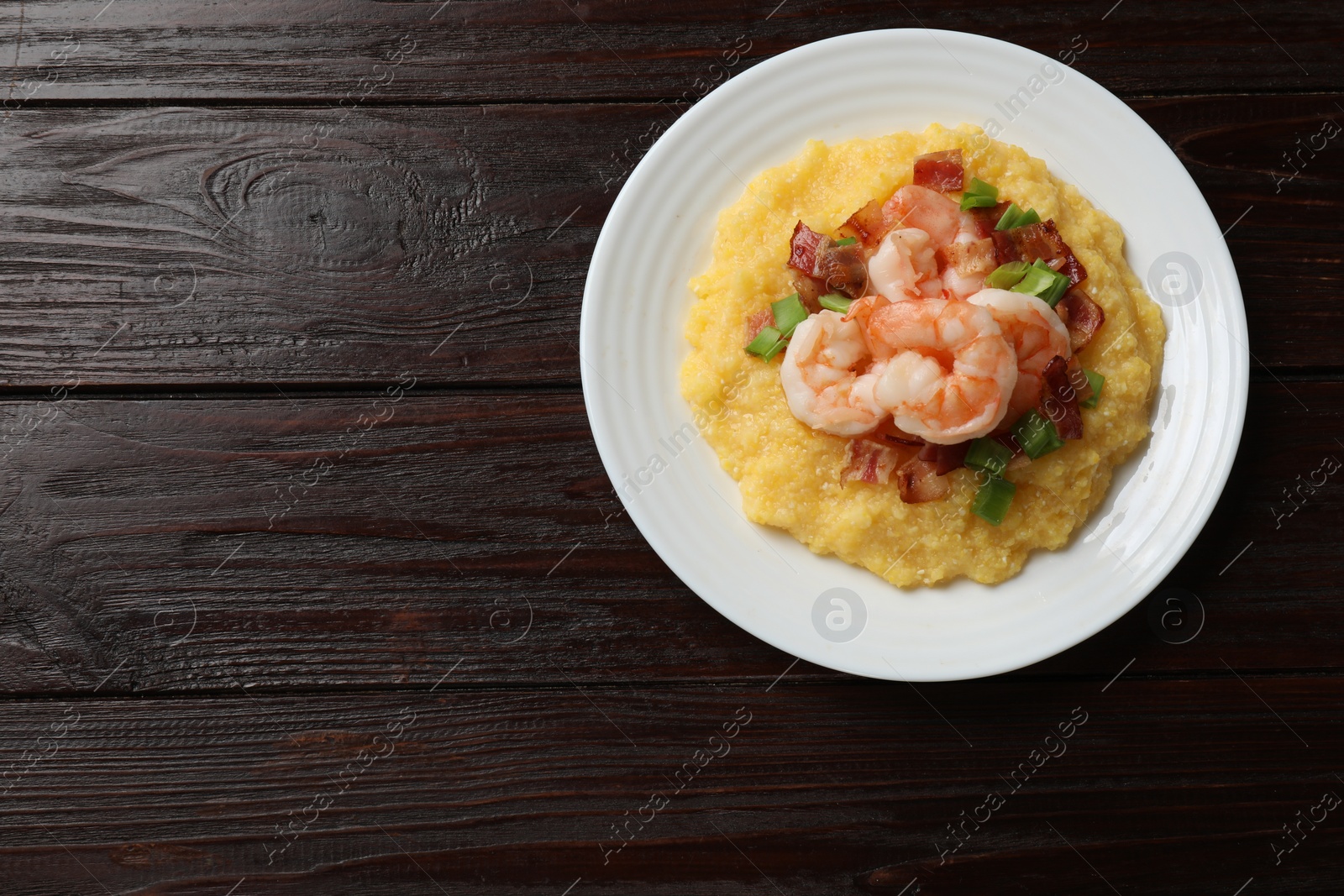 Photo of Plate with fresh tasty shrimps, bacon, grits and green onion on dark wooden table, top view. Space for text