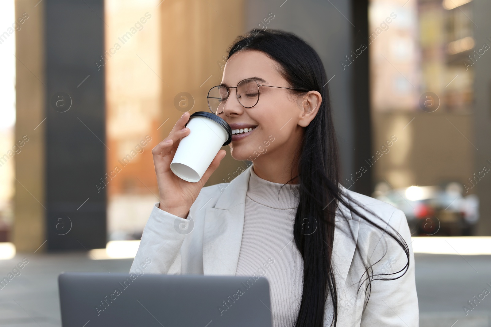 Photo of Happy young woman with cup of coffee using modern laptop outdoors