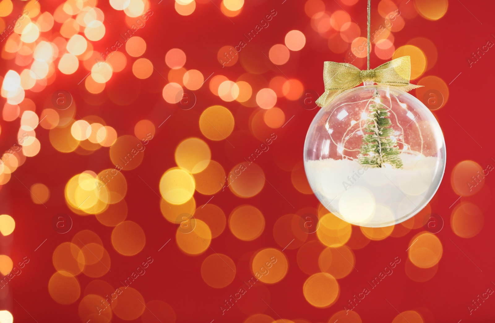 Image of Beautiful transparent Christmas ornament with small fir tree, fairy lights and snow hanging against red background, bokeh effect. Space for text