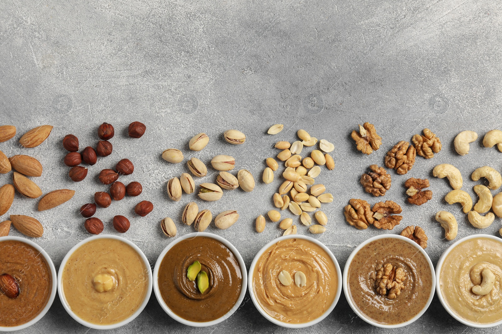 Photo of Tasty nut butters in bowls and raw nuts on light grey background, flat lay