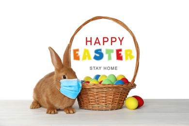 Image of Text Happy Easter Stay Home and cute bunny in protective mask on white table. Holiday during Covid-19 pandemic