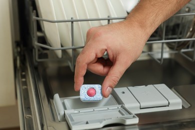 Photo of Man putting detergent tablet into open dishwasher indoors, closeup