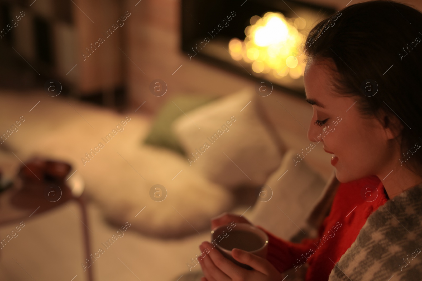 Photo of Young woman drinking coffee near decorative fireplace at home. Winter season