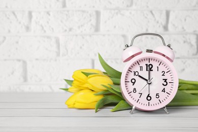 Photo of Pink alarm clock and beautiful tulips on white wooden table against brick wall, space for text. Spring time