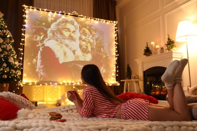 MYKOLAIV, UKRAINE - DECEMBER 24, 2020: Woman watching The Christmas Chronicles movie via video projector in room. Cozy winter holidays atmosphere