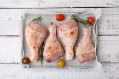 Photo of Raw marinated chicken drumsticks, rosemary and tomatoes in baking dish on white wooden table, top view