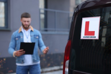 Photo of Instructor with clipboard near car outdoors, selective focus on L-plate. Driving school