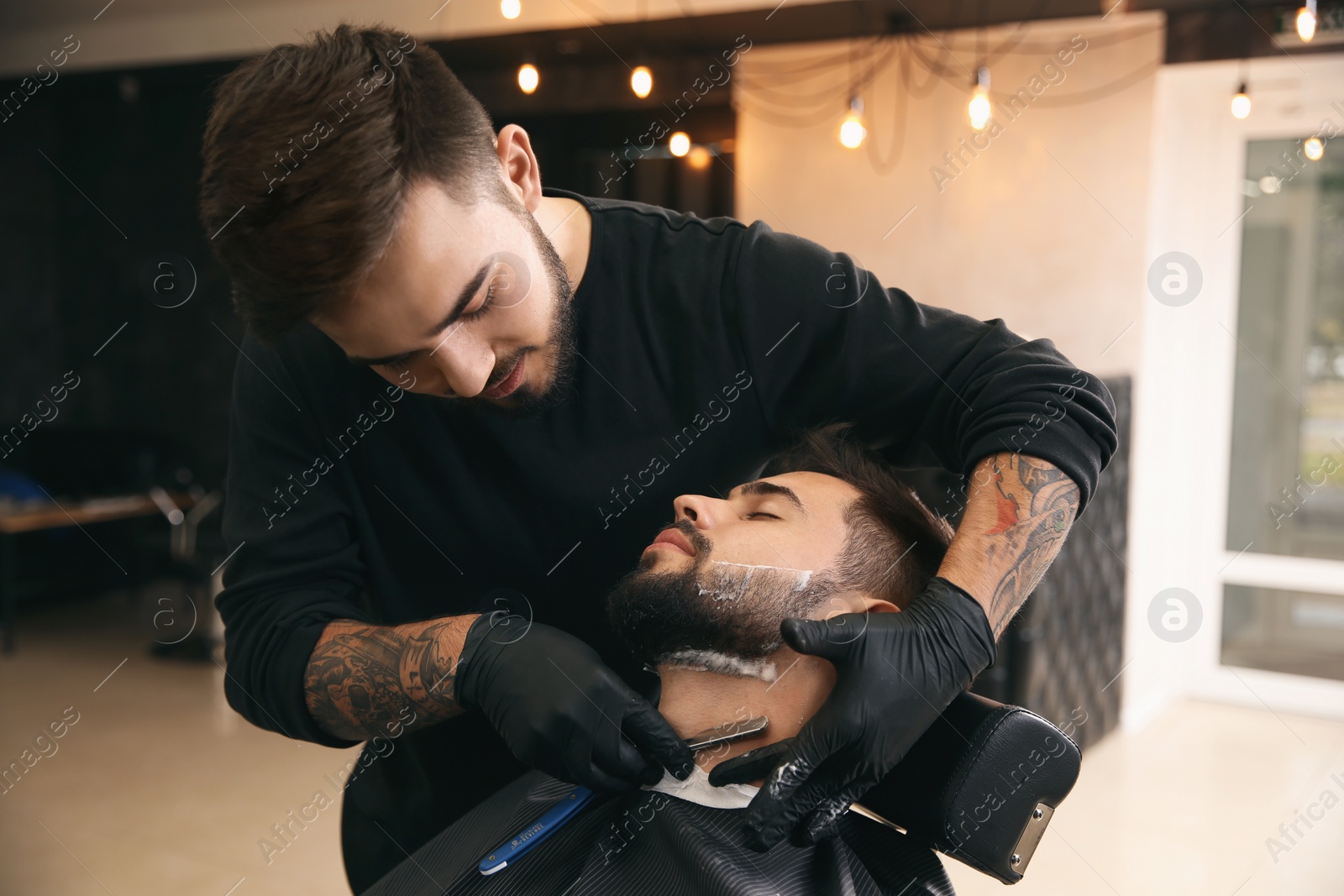 Photo of Professional hairdresser shaving client with straight razor in barbershop