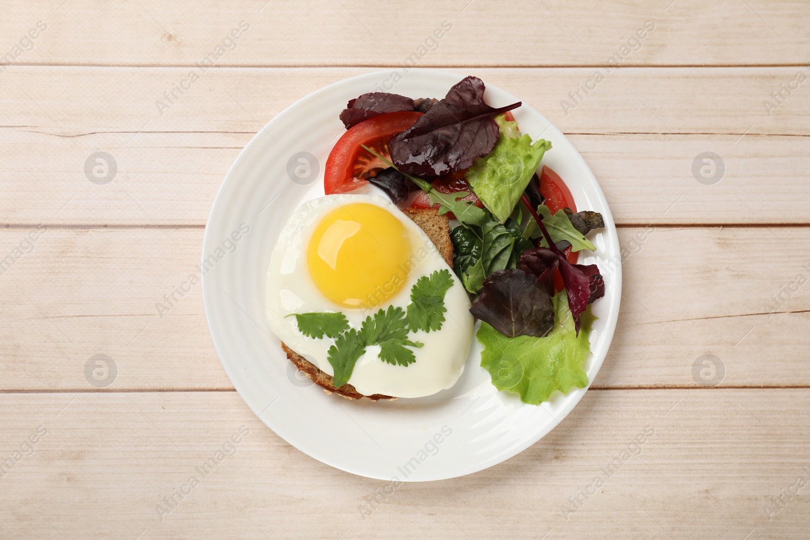 Photo of Plate with tasty fried egg, slice of bread and salad on light wooden table, top view