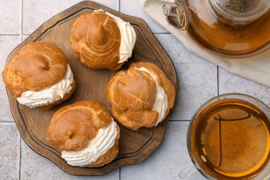 Photo of Delicious profiteroles filled with cream and tea on white tiled table, flat lay