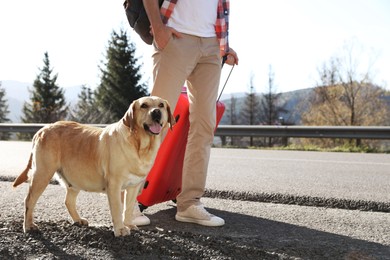 Photo of Man with red suitcase and adorable dog near road outdoors, closeup. Traveling with pet