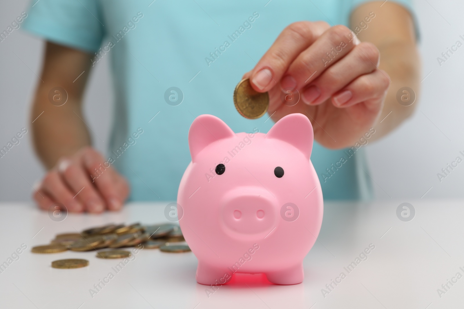 Photo of Woman putting coin into pink piggy bank at white table, closeup