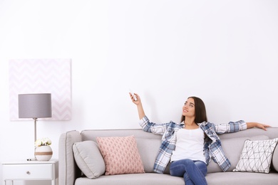 Young woman switching on air conditioner while sitting on sofa near white wall