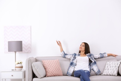 Photo of Young woman switching on air conditioner while sitting on sofa near white wall