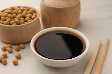 Photo of Soy sauce in bowl, soybeans and chopsticks on white table, closeup