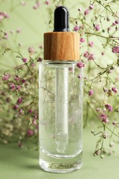 Photo of Bottle of face serum and beautiful flowers on light green background, closeup