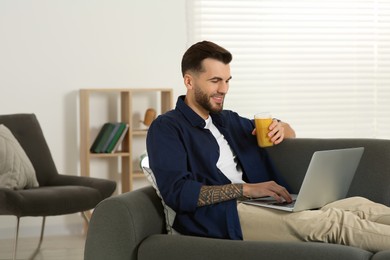 Handsome man with delicious smoothie using laptop on sofa at home
