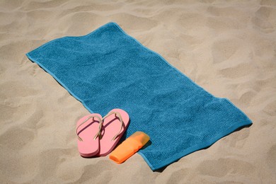 Photo of Soft blue beach towel, pink flip flops and sunscreen on sand. Space for text