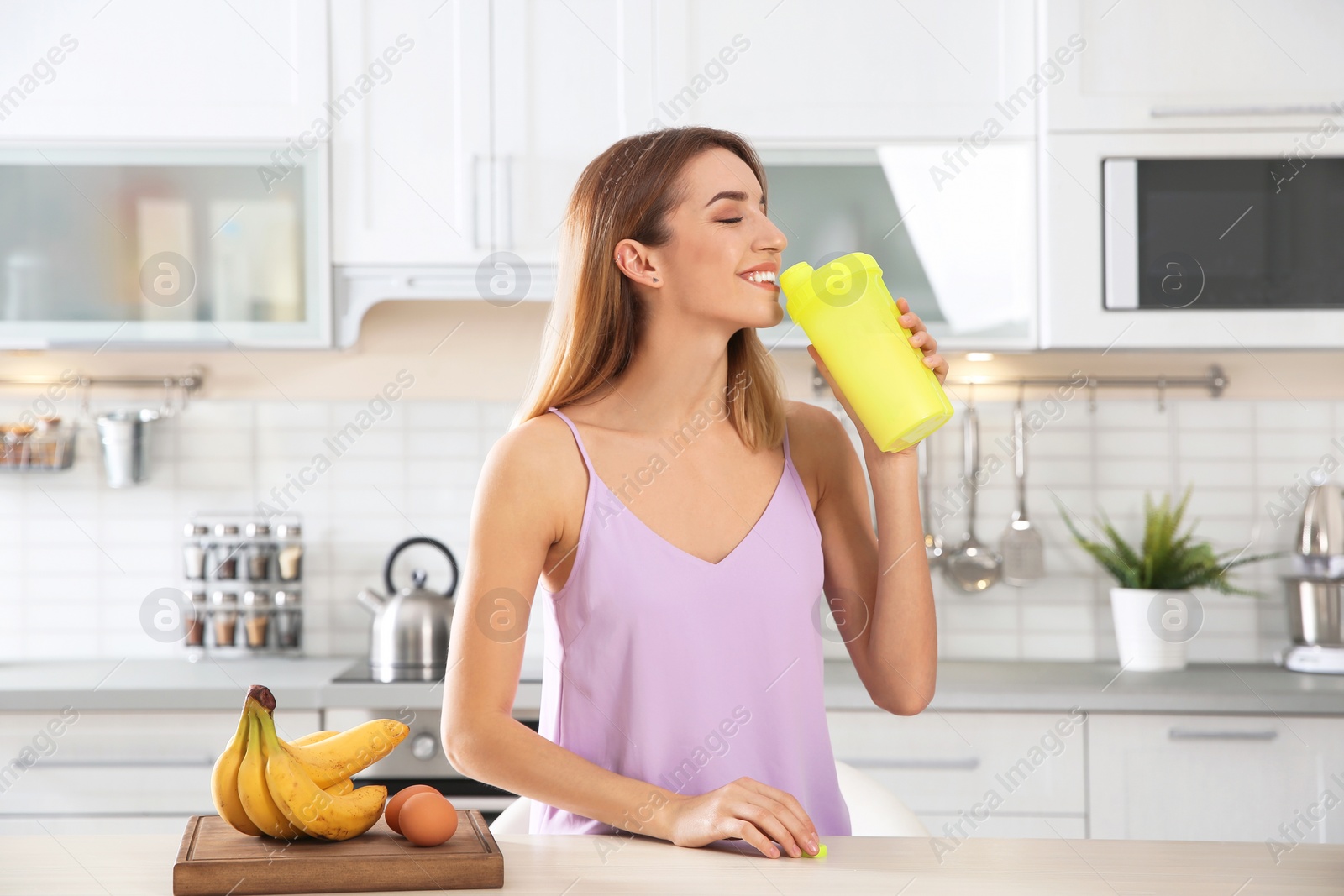 Photo of Young woman drinking protein shake near table with ingredients in kitchen
