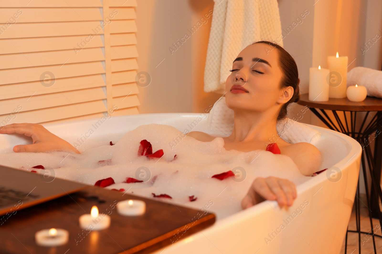 Photo of Woman with laptop taking bath in tub with foam and rose petals indoors