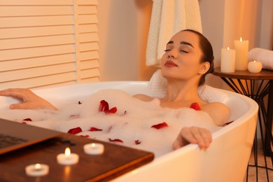 Photo of Woman with laptop taking bath in tub with foam and rose petals indoors