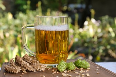 Mug with beer, fresh hops and ears of wheat on wooden table outdoors, space for text