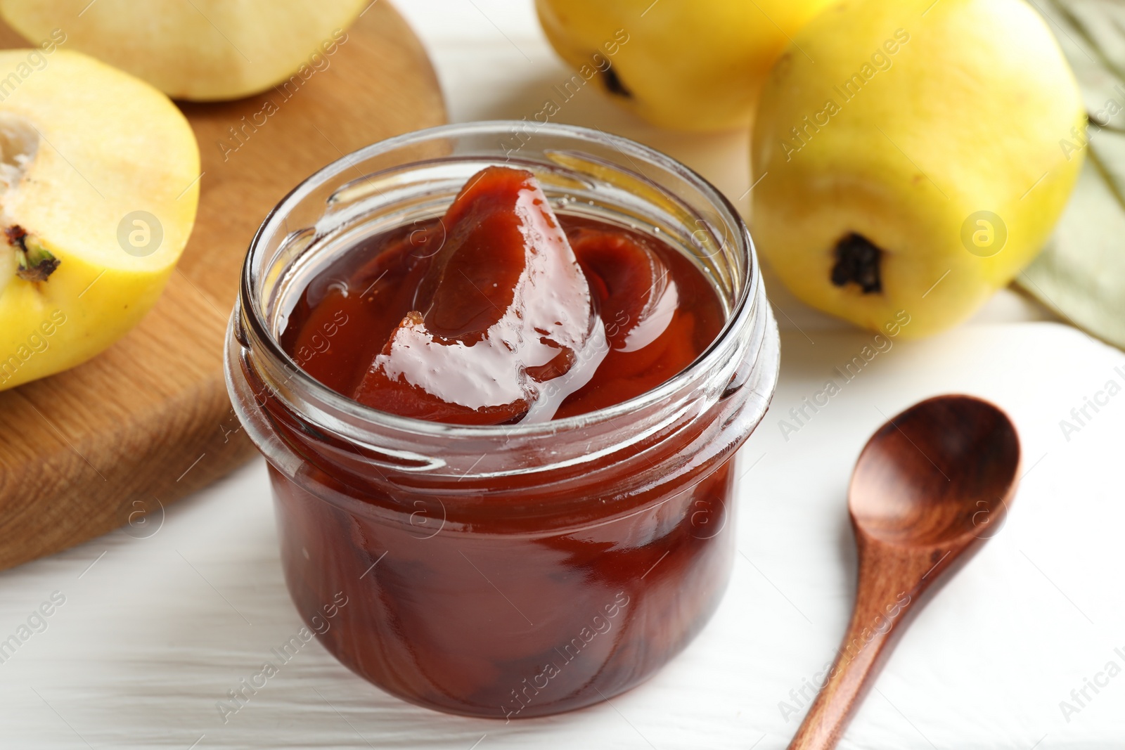 Photo of Tasty homemade quince jam in jar, spoon and fruits on white wooden table, closeup