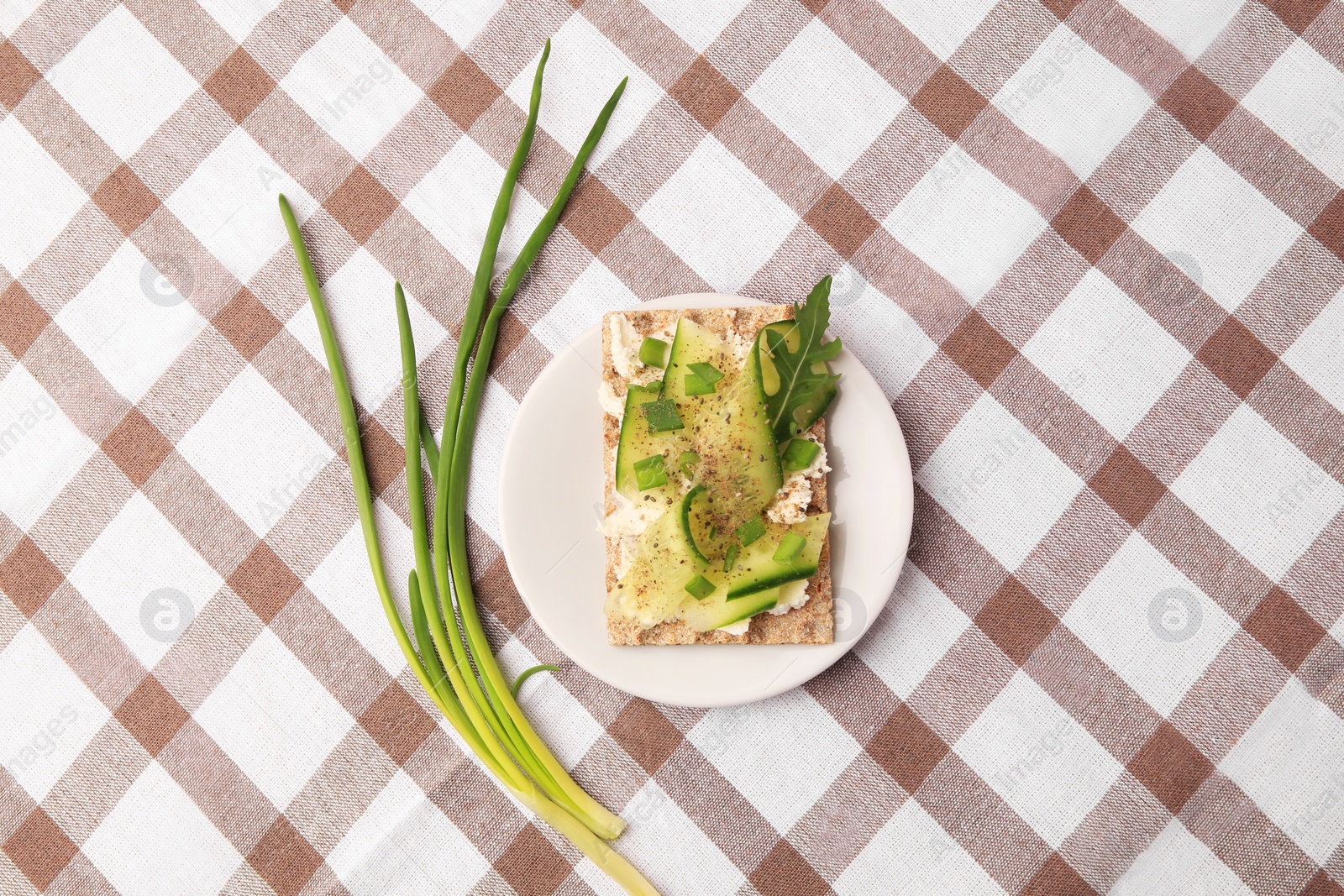 Photo of Fresh crunchy crispbread with cream cheese, cucumber, green onion and arugula on checkered tablecloth, flat lay