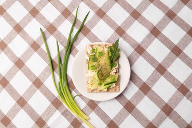 Photo of Fresh crunchy crispbread with cream cheese, cucumber, green onion and arugula on checkered tablecloth, flat lay