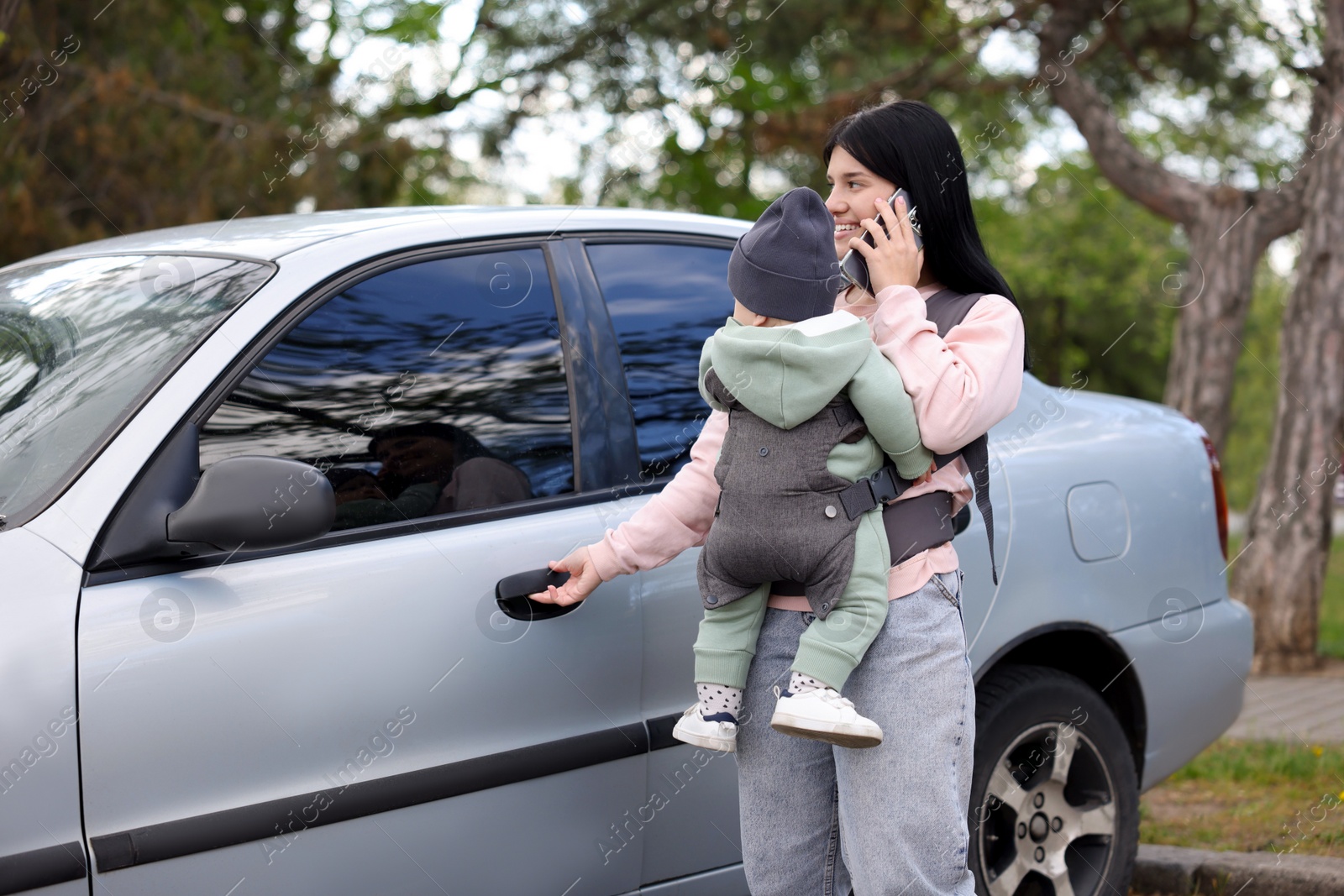 Photo of Mother holding her child in sling (baby carrier) while talking on smartphone near car outdoors